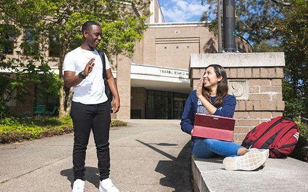 Two students smile and wave to each other outside of the John C. Pace Library