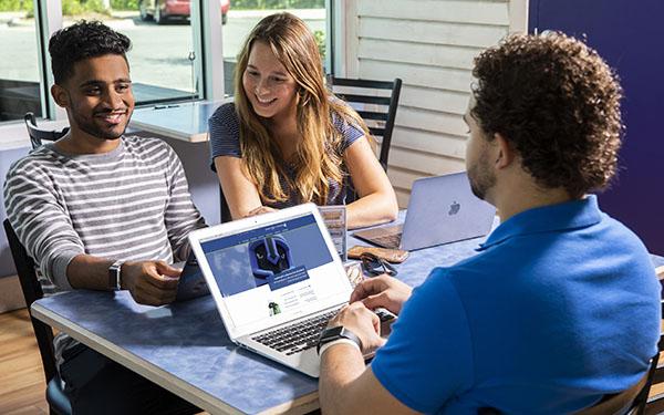 Three students gather at a table with a laptop visiting the UWF website.